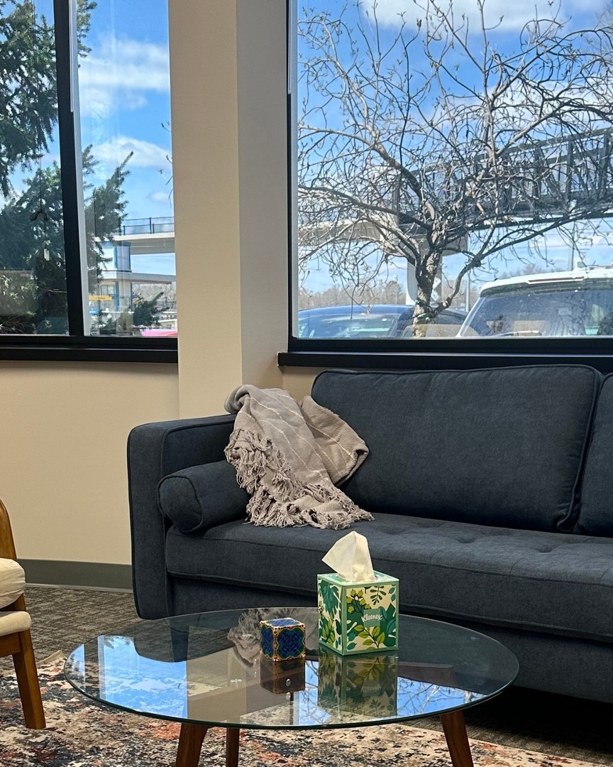 Family and group therapy office at MMHC with a navy sofa, accent chair, and glass coffee table beneath bright windows.