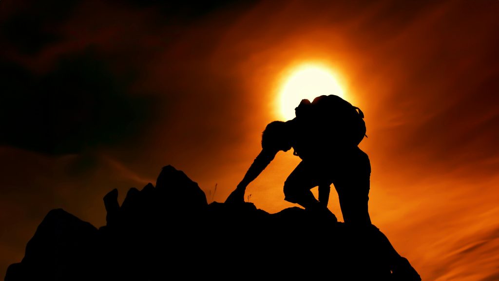 Shadow of a man climbing a mountain with a sun glowing in the background.
